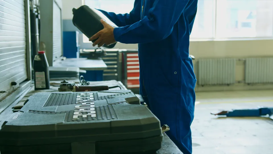 Mechanic in blue coveralls pouring a jug of motor oil at a workbench